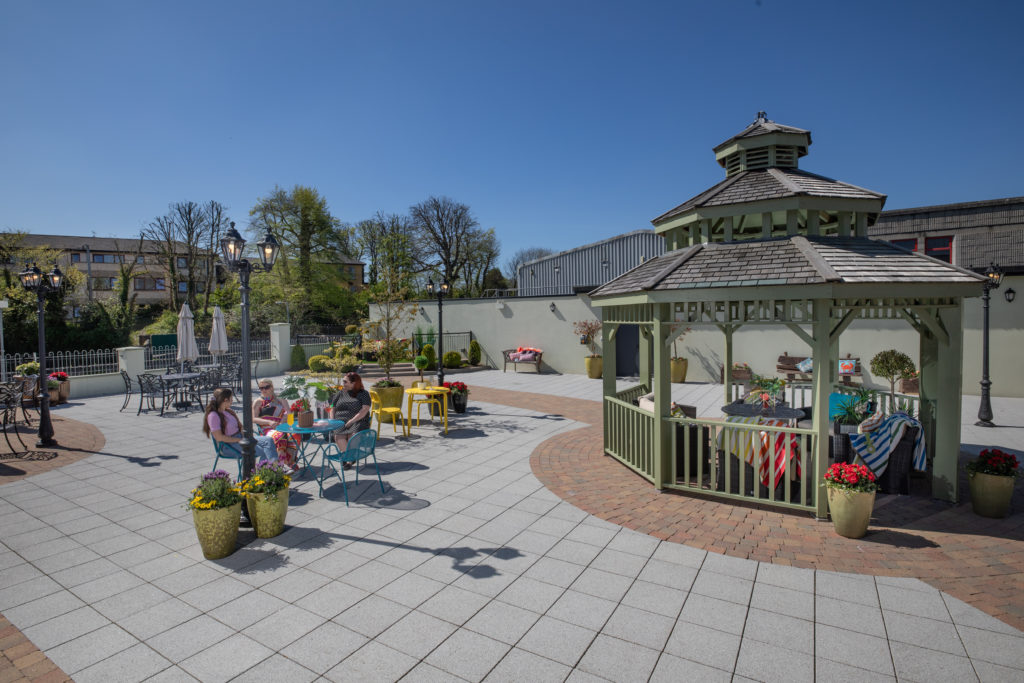 Hotel Courtyard Oasis Guests relax under sunny skies in a charming courtyard with seating, vibrant flowers, and a traditional gazebo.