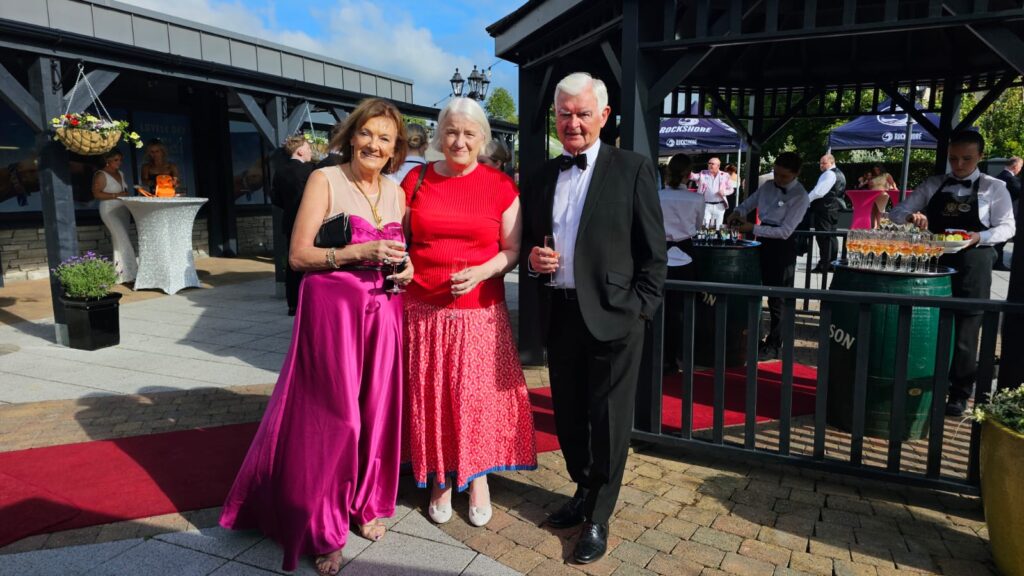 Three adults pose on a red carpet outdoors at a formal event; two women, one in a purple gown and one in a red top and skirt, stand beside a man in a tuxedo. Guests and drinks are visible behind them.