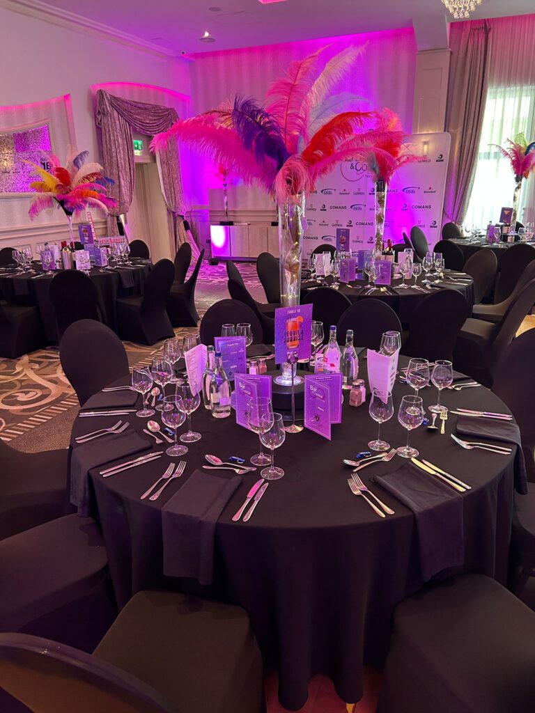Round banquet tables with black tablecloths are set with glassware, silverware, and menus; tall centerpieces with colorful feathers and purple lighting decorate the room.