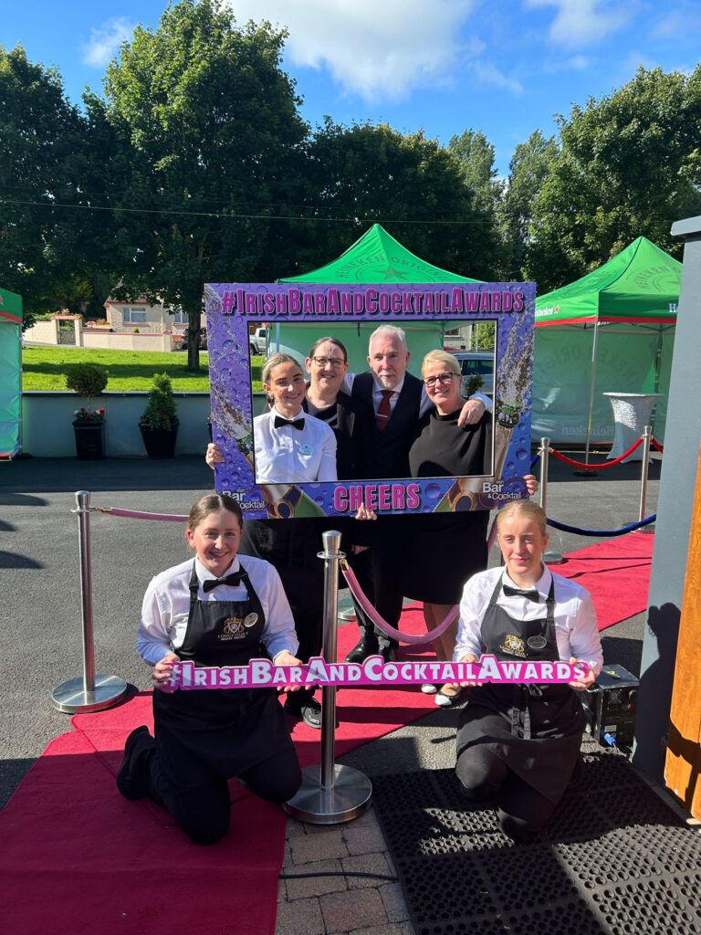 Five people in formal attire pose outdoors at an event, holding large “Irish Bar and Cocktail Awards” frames, with red carpet and green tents visible in the background.