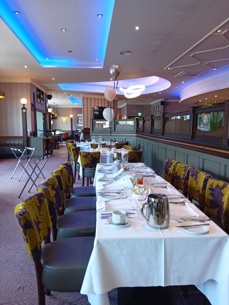 Long dining table set with white tablecloths, menus, and silverware in a modern restaurant with purple and yellow chairs and blue ceiling lights. Balloons decorate the far end of the table.