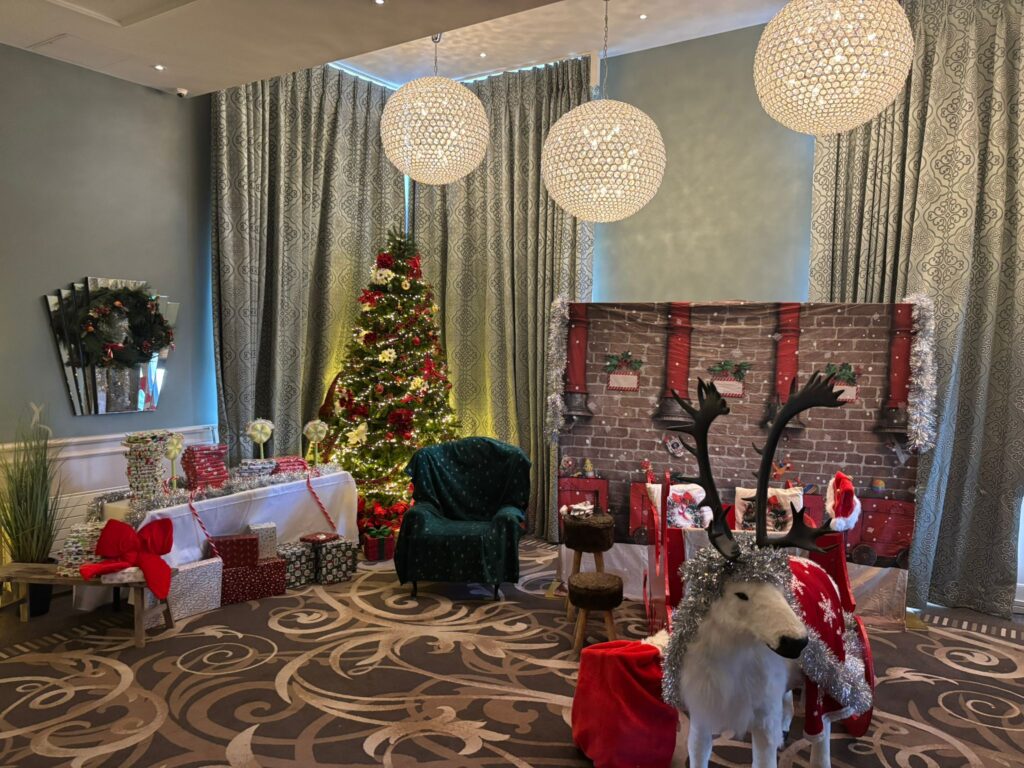 A festive room with a decorated Christmas tree, wrapped gifts, a faux fireplace backdrop, green armchair, and a white reindeer statue with antlers stands in the foreground.