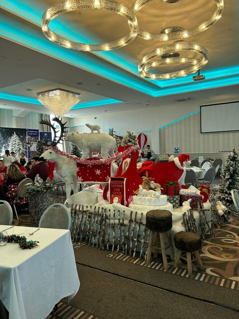 A decorated banquet hall with holiday-themed displays, including a red sleigh, reindeer figures, polar bears, and artificial snow, surrounded by dining tables and festive lights.
