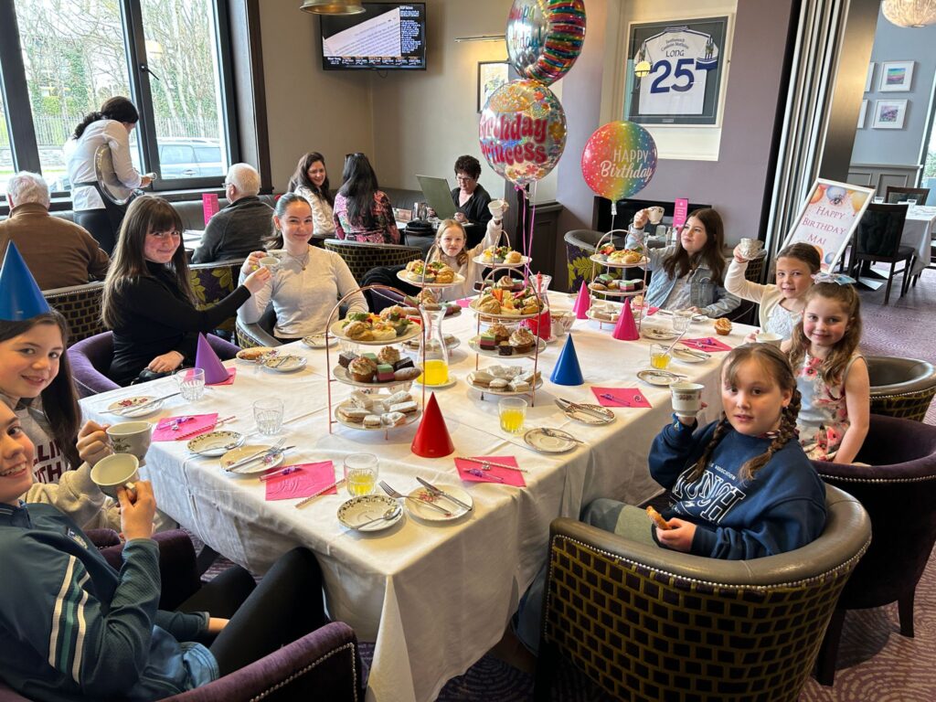 A group of children sit around a table set with afternoon tea, party hats, and birthday balloons in a restaurant.