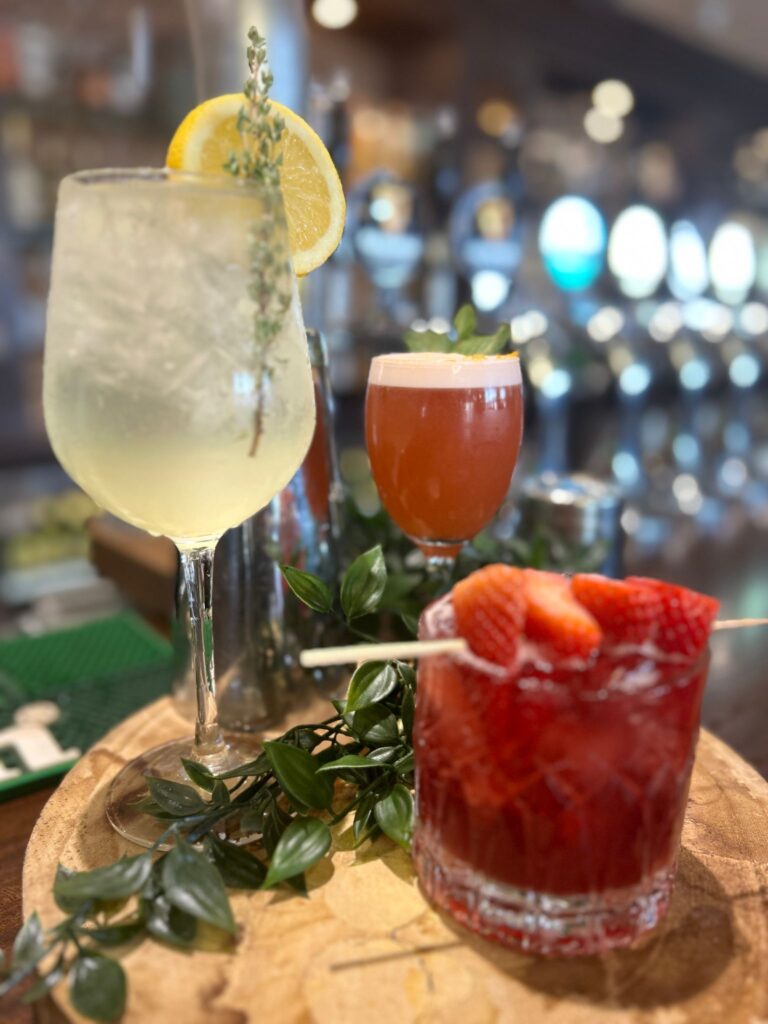 Three cocktails on a wooden tray: a clear drink with lemon and thyme, a red drink with foam and a leaf, and a glass with strawberries, in a bar setting.