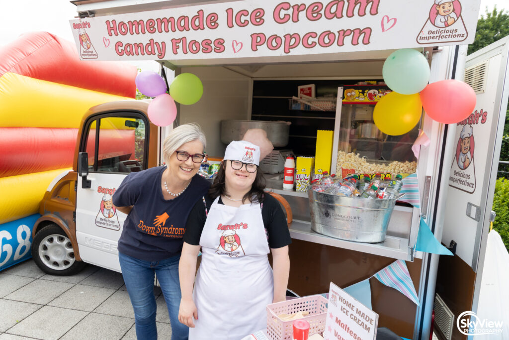 Two women stand in front of an ice cream, candy floss, and popcorn stall decorated with balloons. One wears an apron and hat, and the other stands beside her, smiling.