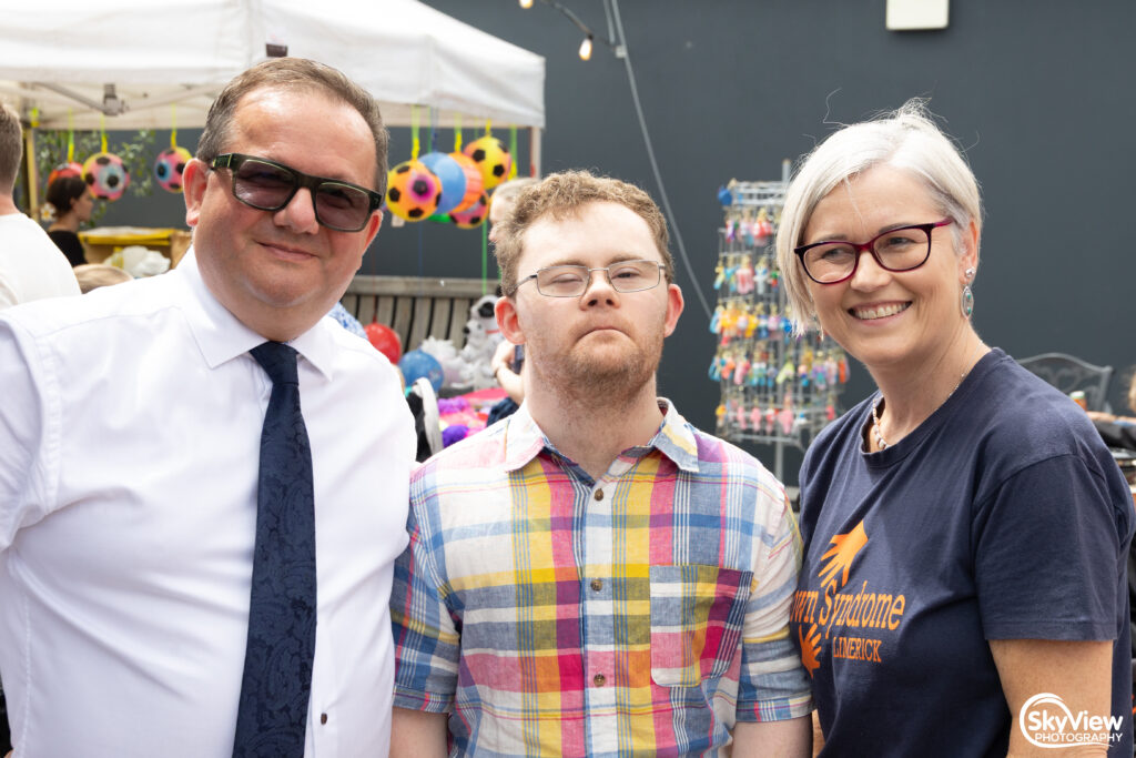 Three adults stand together outdoors at a market or fair, smiling at the camera. Colorful toys and tents are visible in the background.